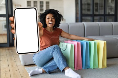 Photo for Shopping App. Happy Young Black Woman Demonstrating Blank Smartphone At Camera While Sitting On Couch Next To Shopping Bags, Smiling African American Lady Recommending Mobile Application, Mockup - Royalty Free Image