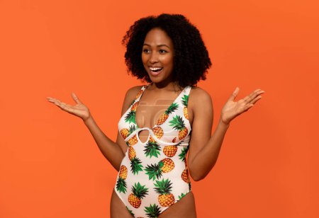Photo for Excited amazed happy millennial african american beautiful curly woman in swimwear gesturing raising hand up and laughing over orange studio background, looking at copy space for ad - Royalty Free Image