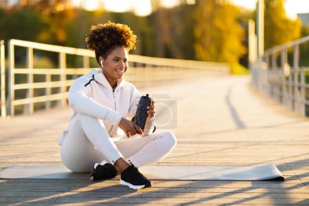Foto de Mujer negra deportiva escuchando música y agua potable mientras se relaja después del entrenamiento al aire libre, sonriendo a una joven afroamericana haciendo ejercicio físico afuera, sentada en una esterilla de yoga y mirando hacia otro lado - Imagen libre de derechos