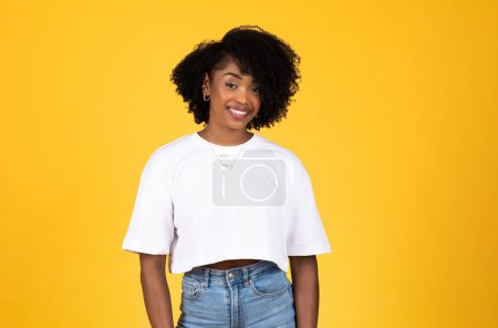 Photo for Smiling millennial african american curly woman in white t-shirt looking at camera, isolated on yellow studio background. Student casual lifestyle, ad and offer, study and fashion - Royalty Free Image
