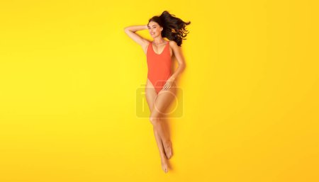 Téléchargez les photos : Protection UV d'été. Happy Young Woman In Orange Maillot de bain une pièce posant sur fond jaune studio. Bain de soleil et vacances Concept de détente. Vue du dessus, Panorama - en image libre de droit