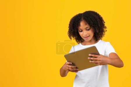 Téléchargez les photos : Joyeux adolescent frisé afro-américaine fille en t-shirt blanc joue jeu en ligne sur tablette, regarder la leçon vidéo à distance, isolé sur fond de studio jaune. Site Web pour l'éducation, l'étude et le chat - en image libre de droit