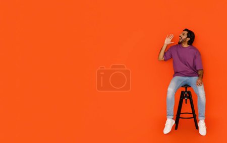 Photo for Cool happy handsome young eastern guy wearing stylish casual outfit sitting on chair on orange studio background, shouting towards copy space for advertisement, making announcement, full length shot - Royalty Free Image