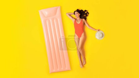 Téléchargez les photos : Summer Joy. Joyeux jeune femme en maillot de bain orange couché près du Lilo gonflable, bronzant et relaxant ayant piscine partie sur fond jaune studio, souriant à la caméra. Vue d'en haut, Panorama - en image libre de droit