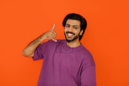 Téléchargez les photos : Communication. joyeux beau barbu confiant millénariste indien gars en tenue décontractée violet t-shirt souriant à la caméra et montrant appelez-moi geste, posant seul sur fond de studio orange - en image libre de droit