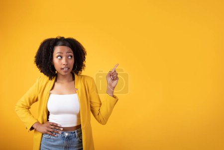 Téléchargez les photos : Portrait de jeune femme noire pointant vers l'espace de copie, femelle debout sur fond de studio jaune, démontrant un endroit vide pour la publicité ou votre texte - en image libre de droit
