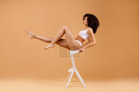 Téléchargez les photos : Jeune femme latine mince en sous-vêtements blancs assis sur une chaise blanche sur fond de studio beige, vue latérale, espace libre. Forme corporelle parfaite et concept de beauté naturelle - en image libre de droit