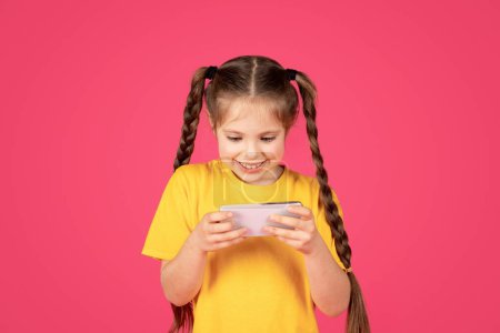 Photo for Cheerful preteen girl playing games on smartphone while standing on pink studio background, cute female child looking at mobile phone screen and smiling, enjoying online gaming, copy space - Royalty Free Image