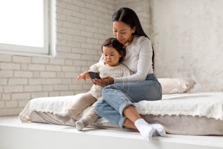 Téléchargez les photos : Mère chinoise et tout-petit bébé fille en utilisant le téléphone mobile Jouer à un jeu éducatif en ligne et regarder des dessins animés assis dans une chambre confortable à l'intérieur. Concept d'utilisation de bébé et de gadget - en image libre de droit