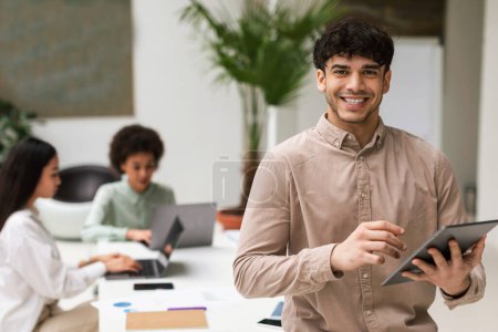 Photo for Digital Business Offer. Joyful Middle Eastern Businessman With Tablet Standing At Corporate Meeting In Modern Office, Posing In Front Of Colleagues And Smiling To Camera. Career And Leadership - Royalty Free Image