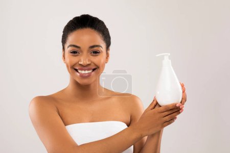 Photo for Happy smiling beautiful young african american woman wrapped in bath towel holding white bottle with beauty product, recommending shower gel, body lotion, shampoo, pastel studio background - Royalty Free Image