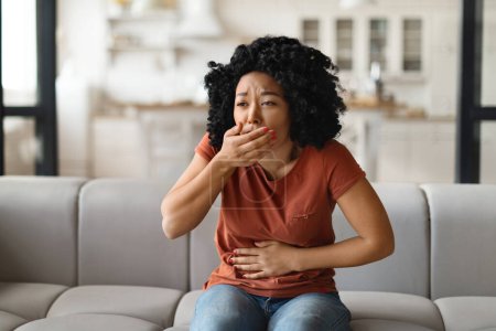 Young Black Woman Suffering From Nausea At Home, Millennial African American Female Having Morning Sickness, Ill Lady Sitting On Couch In Living Room, Covering Mouth And Touching Belly