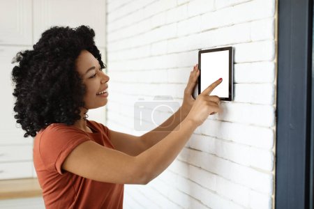 Photo for Innovative Technology. Black Woman Using Digital Tablet With Empty Screen On Wall With Smart Home Control System App, African American Lady Adjusting House Automation Processes And Settings, Mockup - Royalty Free Image