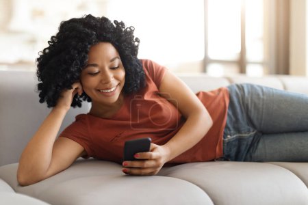 Foto de Mujer negra joven usando Smartphone mientras descansa en el sofá en casa, Hermosa mujer afroamericana acostada en el sofá con teléfono móvil, Mensajería con amigos o navegar por la nueva aplicación, Primer plano - Imagen libre de derechos