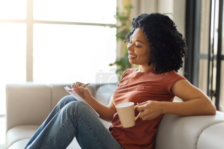 Téléchargez les photos : Joyeux jeune femme noire se détendre à la maison, boire du café et écrire dans un bloc-notes, sourire femme afro-américaine assis sur le canapé avec une tasse de thé, prendre des notes, remplir un journal ou faire une liste de contrôle - en image libre de droit