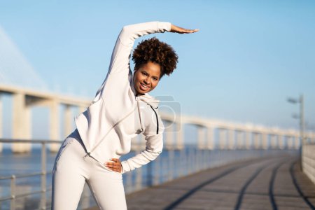 Téléchargez les photos : Femme noire sportive faisant de l'exercice de flexion latérale pendant l'entraînement en plein air, Jeune femme afro-américaine de jogging se réchauffant avant la course du matin, s'exerçant sur une jetée en bois près de la mer, Copier l'espace - en image libre de droit
