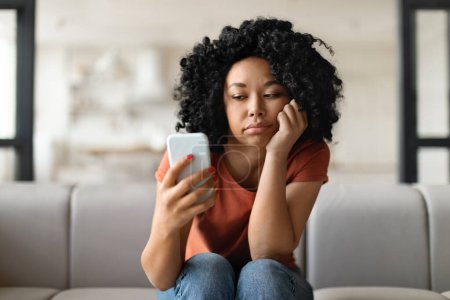 Photo for Bored Young Black Female Looking At Smartphone Screen While Sitting On Couch At Home, Pensive African American Woman Holding Mobile Phone, Waiting For Important Call Or Sms, Copy Space - Royalty Free Image