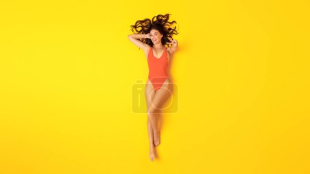 Téléchargez les photos : Summer Joy. Femme gaie en orange maillot de bain une pièce gestuelle signe de victoire souriant à la caméra allongé sur fond jaune, vue du dessus. Vacances Loisirs, bains de soleil et détente - en image libre de droit