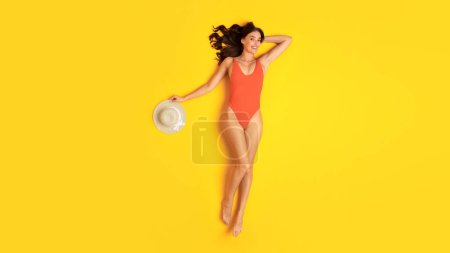 Téléchargez les photos : Heure d'été. Femme mince gaie portant un maillot de bain orange et un chapeau, allongé souriant à la caméra posant sur fond jaune studio. Publicité de fête de vacances et de piscine. Panorama - en image libre de droit
