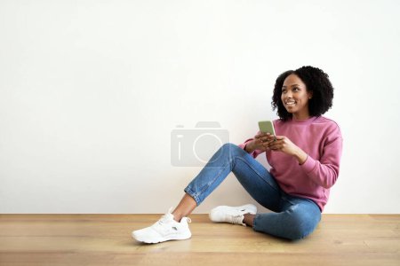 Téléchargez les photos : Glad millénaire afro-américaine frisée femme tapant sur smartphone, assis sur le sol à l'intérieur de la chambre, regarder l'espace libre sur le mur blanc. Chat, appel vidéo à distance, travail et étude à la maison, annonce, offre - en image libre de droit
