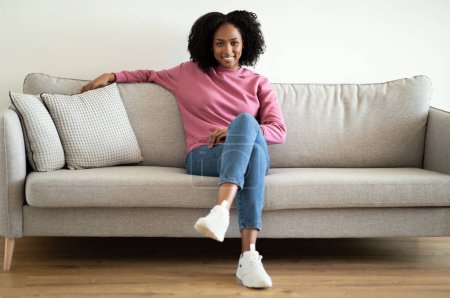 Téléchargez les photos : Souriant confiant millénariste afro-américain dame bouclée dans casual assis sur canapé, profiter du confort et du temps libre dans l'intérieur du salon. Repos, détente, pause et style de vie à la maison - en image libre de droit