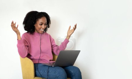 Foto de Sorprendido alegre millennial afroamericano rizado dama en casual se sienta en la silla, levanta las manos, mira a la computadora portátil en fondo blanco de la pared. Sorpresa, trabajo y estudio, venta, videollamada en casa - Imagen libre de derechos