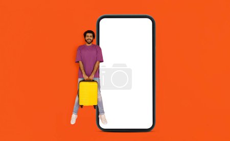 Foto de Aplicación de reserva. Emocionado joven indio guapo feliz en turista casual sosteniendo equipaje amarillo y saltando en el aire por un enorme teléfono inteligente con pantalla blanca en blanco, aislado sobre fondo naranja, maqueta - Imagen libre de derechos
