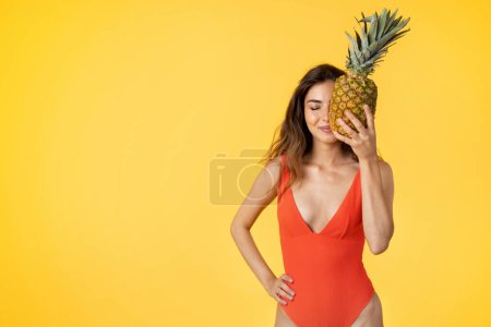 Photo for Happy millennial mixed race lady in swimsuit hold pineapple, has fun, enjoy fruit, isolated on yellow studio background. Vacation trip in summer, diet at holiday, resort at summer - Royalty Free Image