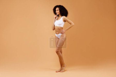 Téléchargez les photos : Jeune femme latine mince en sous-vêtements blancs posant isolée sur fond studio beige, prise de vue pleine longueur, espace libre. Jeune femme avec ajustement corps mince regardant de côté à l'espace de copie - en image libre de droit