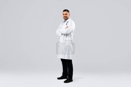 Photo for Cheerful middle aged man doctor posing with folded arms looking at camera standing on light background, wearing white uniform, coat and stethoscope, full body length, free space - Royalty Free Image