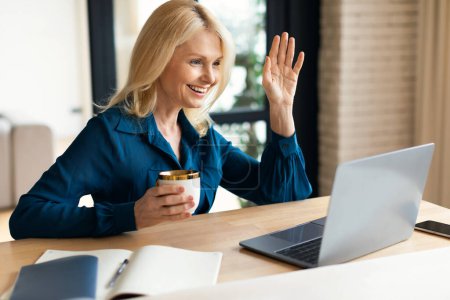 Photo for Happy mature businesswoman drinking coffee and having business meeting, waving hand at laptop, sitting at table at home. Greeting, hi gesture, remote conference and coffee break - Royalty Free Image