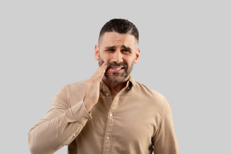 Photo for Unhappy middle aged businessman suffering from tooth ache, having gum problems, touching his cheek and looking at camera, standing on light studio background. Male suffering from dental pain - Royalty Free Image