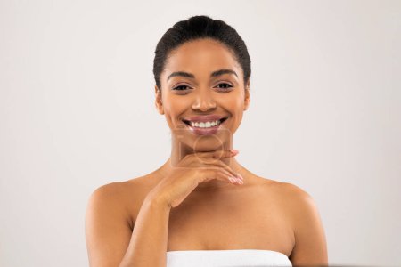 Téléchargez les photos : Portrait de heureuse jeune femme afro-américaine attrayante enveloppée dans une serviette de bain touchant sa peau lisse et soyeuse et souriante, obtenir un traitement spa facial, isolé sur fond de couleur pastel - en image libre de droit