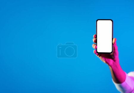 Téléchargez les photos : Online offer, mobile app. Cropped of african american man hand holding modern cell phone with white blank screen on blue studio background in neon light, mockup for advertisement, copy space - en image libre de droit