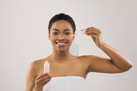 Téléchargez les photos : Jolie jeune femme noire souriante enveloppée dans une serviette tenant une petite bouteille avec un produit de beauté, utilisant un sérum hydratant anti-âge ou une huile nourrissante pour une peau jeune, un fond de studio - en image libre de droit