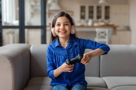 Foto de Gadgets Pasatiempo. Chica alegre jugando videojuegos en línea con auriculares, sosteniendo el controlador del mando del mando sentado en el sofá en casa el fin de semana. Diversión digital, Internet Entretenimiento Actividades - Imagen libre de derechos
