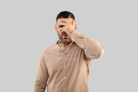 Photo for Curious middle aged man peeking covering eyes and looking at camera through fingers posing over light studio background. Curiosity concept, advertisement banner - Royalty Free Image