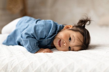 Téléchargez les photos : Charme de bébé. Mignon japonais bébé fille posant couché sur l'estomac souriant à la caméra dans une chambre confortable à la maison. Prise de vue d'un adorable tout-petit enfant portant un jean Vêtements à l'intérieur. Mode et style enfants - en image libre de droit