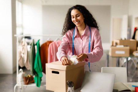 Photo for Fashion Business. Cheerful Lady Clothing Designer Packing Trendy Clothes In Cardboard Box, Getting Ready Order For Client Working In Store Office Indoors. Ecommerce, Successful Entrepreneurship - Royalty Free Image