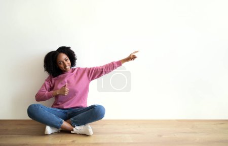 Téléchargez les photos : Heureuse dame frisée afro-américaine millénaire assis sur le sol, pointe du doigt l'espace vide, montrer pouce vers le haut sur fond de mur blanc. Recommandation vente, conseil annonce, offre à domicile - en image libre de droit