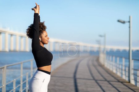 Téléchargez les photos : Belle femme noire athlétique étirant les bras avant l'entraînement de remise en forme en plein air, souriante jeune afro-américaine en vêtements de sport debout sur une jetée en bois près de la mer, se réchauffant pour le jogging - en image libre de droit