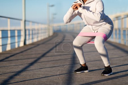Téléchargez les photos : Entraînement de fitness. Entraînement de jeune femme noire avec bande de boucle de résistance à l'extérieur, Femme afro-américaine méconnaissable faisant de l'exercice sur une jetée en bois près de la mer, faisant des activités de perte de poids, Copier l'espace - en image libre de droit