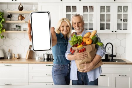 Téléchargez les photos : Couple de personnes âgées tenant un sac plein d'épicerie et montrant smartphone vide avec écran vide tout en restant dans l'intérieur de la cuisine, conjoints âgés en utilisant l'application mobile pour la livraison d'épicerie, maquette - en image libre de droit
