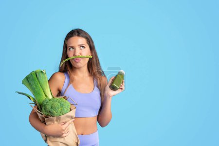 Photo for Positive millennial caucasian slim lady in sportswear with vegetable bag, show bottle of smoothie, has fun, isolated on blue studio background. Sports, slimming, diet and body care, ad, offer - Royalty Free Image