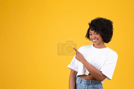 Téléchargez les photos : Femme frisée afro-américaine millénaire souriante en t-shirt blanc pointant du doigt l'espace vide, isolée sur fond de studio jaune. Annonce et offre, conseils et recommandations - en image libre de droit