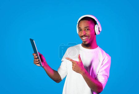 Téléchargez les photos : Beau jeune homme noir barbu positif portant une chemise blanche pointant vers la tablette numérique dans sa main et souriant, utilisant un casque sans fil, écoutant de la musique via une application mobile, fond bleu - en image libre de droit