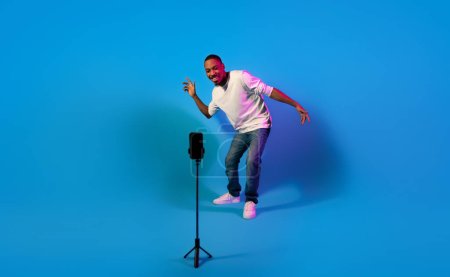 Téléchargez les photos : Célèbre influenceur afro-américain cool élégant millennial guy enregistrement de contenu pour les abonnés, en utilisant un téléphone portable sur trépied, danser et sourire sur fond bleu dans la lumière au néon, pleine longueur - en image libre de droit