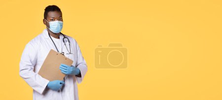 Photo for Medical Staff. Confident African American Physician Doctor Man Posing Wearing Face Mask, White Coat And Rubber Gloves, Holding Clipboard Standing On Yellow Studio Background. Panorama, Copy Space - Royalty Free Image