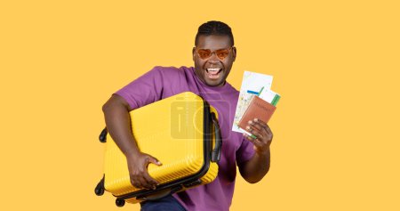Foto de Oferta de vacaciones de verano. Hombre turista negro alegre con maleta mostrando sus boletos de viaje y mapa, posando usando gafas de sol en el fondo del estudio amarillo. Retrato del viajero. Panorama - Imagen libre de derechos