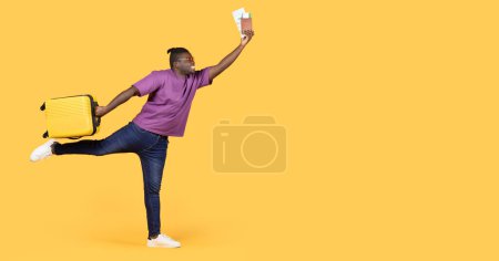 Téléchargez les photos : Les vacances, enfin. Joyeux voyageur afro-américain posant avec une valise de voyage et des billets, se précipitant sur son vol regardant à côté de l'espace de copie sur fond jaune studio. Panorama - en image libre de droit